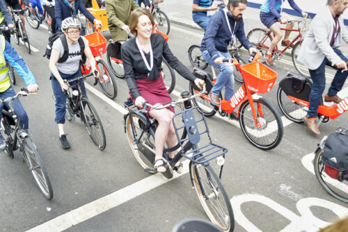 Riders, some on orange hire bikes, cycling together across a road junction on the 2019 Parliamentary Bike Ride