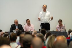 A man standing to speak beside three panellists in front of a seated audience at the 2023 Parliamentary Bike Ride event