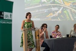 A woman in a floral dress speaking at a lectern beside seated panellists at the 2023 Parliamentary Bike Ride event