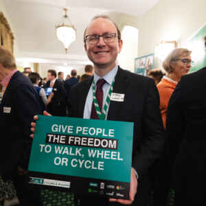 A smiling man in glasses holding a sign reading Give people the freedom to walk, wheel or cycle at the parliamentary reception