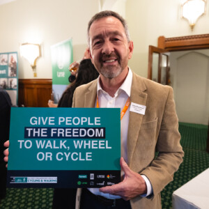 A smiling man in a tan jacket holding a Give people the freedom to walk, wheel or cycle sign at the parliamentary reception