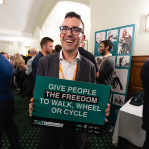 A man in glasses laughing as he holds a Give people the freedom to walk, wheel or cycle sign at the parliamentary reception