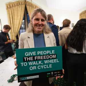 A smiling woman in a cream jacket holding a Give people the freedom to walk, wheel or cycle sign at the parliamentary reception