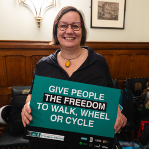 A smiling woman in glasses holding a Give people the freedom to walk, wheel or cycle sign at the parliamentary reception